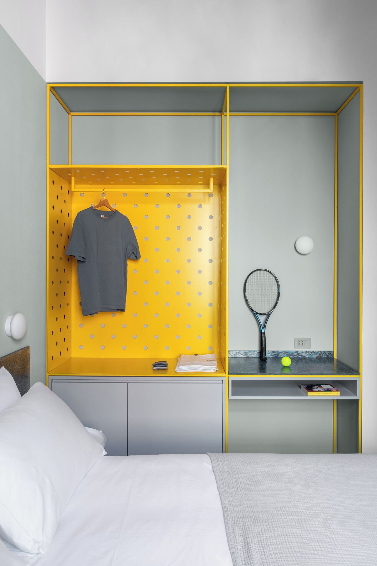 A bedroom with a bed, a wardrobe with a hanging section, a desk with a tennis racket and ball, and a small shelf with a book.
