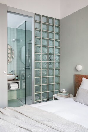 A modern hotel room with a glass partition, a bed, a bedside table with a cup and books, and a bathroom with a glass shower enclosure. -  Ehi Vittorio (Rome)