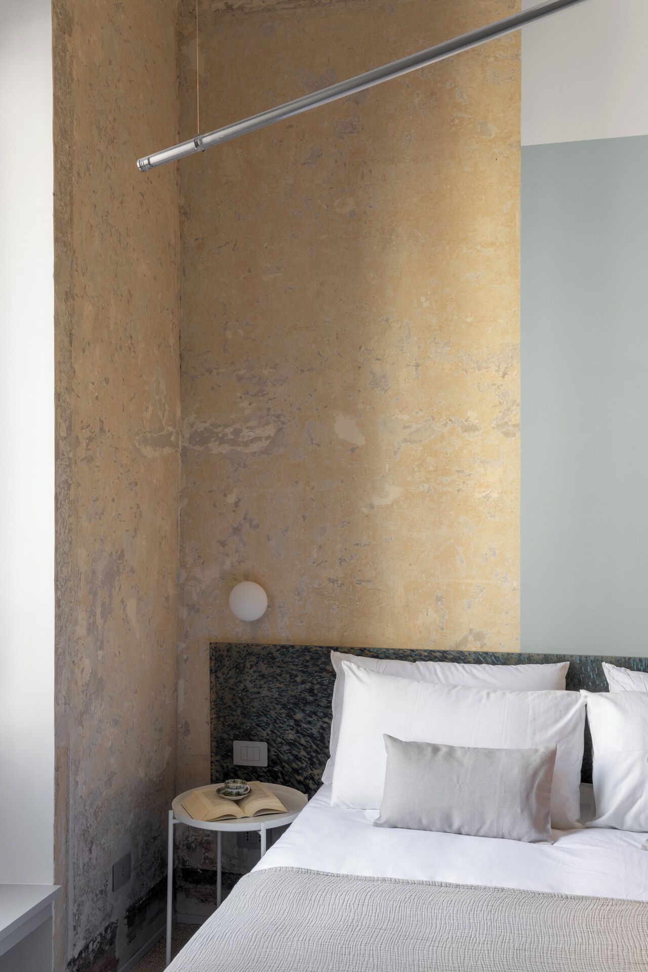 A minimalist bedroom with a bed, pillows, a bedside table with a book, and a wall-mounted light fixture.