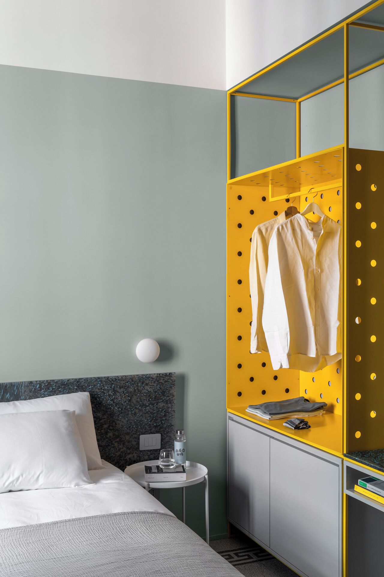 Ein modernes Hotelzimmer mit einem Bett, einem kleinen runden Tisch mit einer Wasserflasche und Gläsern sowie einem gelben Stauraum mit Kleidung.