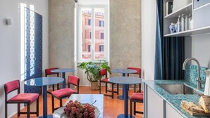 Uma área de refeições compacta, com mesas redondas e cadeiras vermelhas, uma planta em vaso e vista para edifícios através de uma janela.
