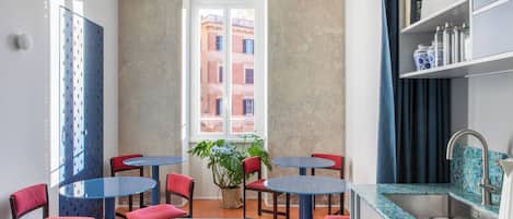 A compact dining area with round tables and red chairs, a potted plant, and a view of buildings through a window.