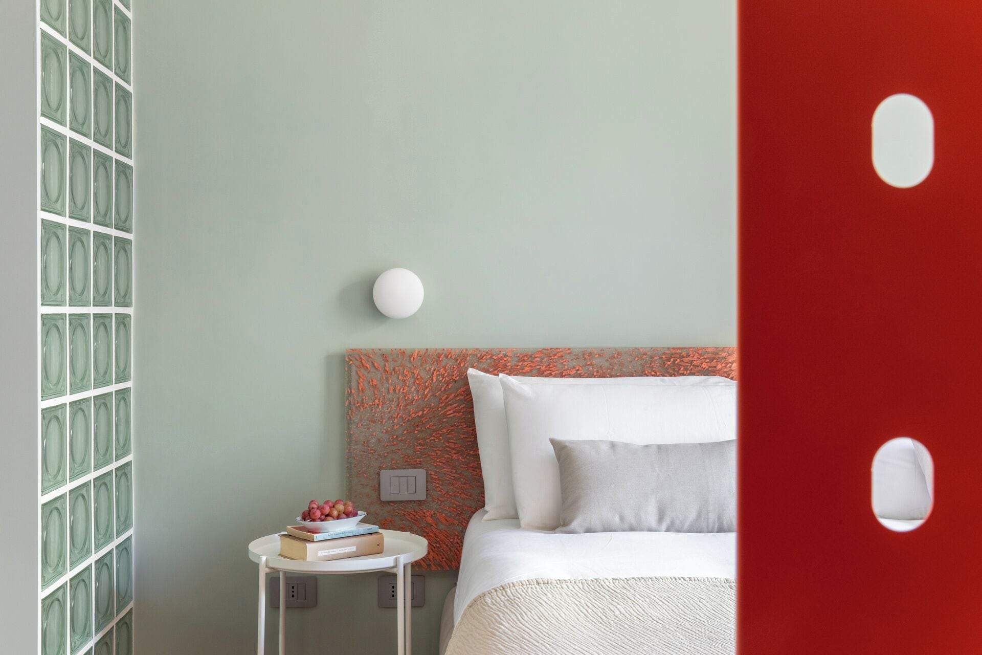 A bedroom with a bed, a bedside table with a bowl of fruit, and a wall with a patterned design.