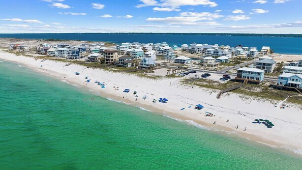 Vue aérienne d’une zone côtière avec une plage, des eaux turquoise et limpides, et une rangée de maisons de plage avec des parasols.
