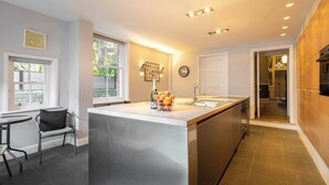 A modern kitchen with a central island, a dining area with a chair, and a view of the backyard through a window. - Host & Stay | Chatsworth Townhouse I (Carlisle)