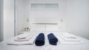 Una cama bien hecha con toallas blancas y azules, una mesita de noche con una radio y una ventana sobre la cama.