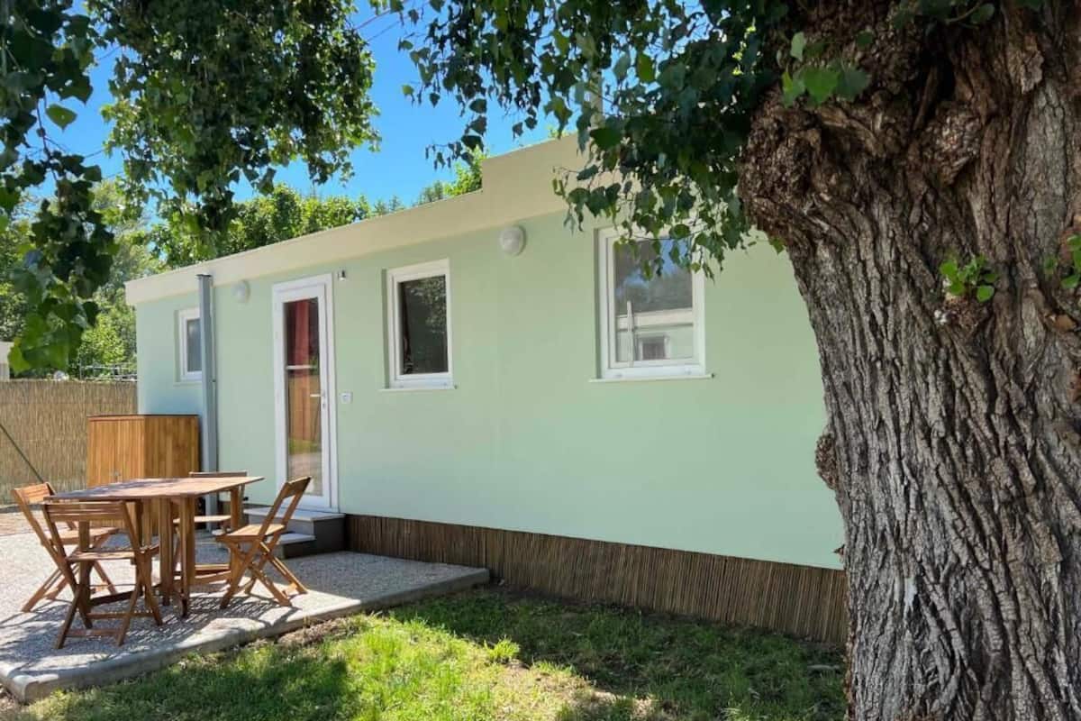 Un bâtiment de couleur vert clair, avec une table et des chaises en bois à l’extérieur, un arbre au premier plan et un ciel dégagé.