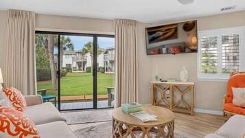 A living room with a large sliding glass door leading to a garden, a ceiling fan, and a flat-screen TV mounted on the wall.