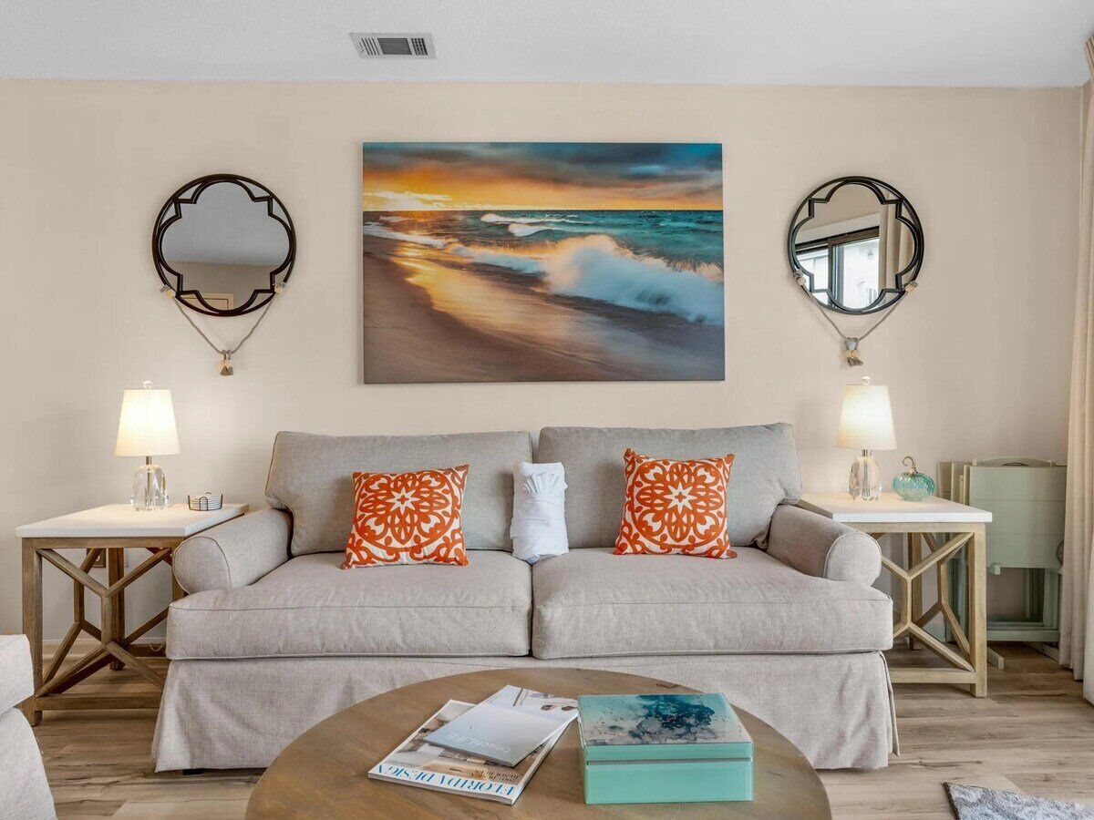 A living room with a beige sofa, two patterned pillows, a coffee table with magazines, and a painting of a sunset over the ocean.