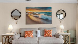 A living room with a beige sofa, two patterned pillows, a coffee table with magazines, and a painting of a sunset over the ocean. - Crystal Cove 28A-Walk to Beach-Sleeps 6 (Miramar Beach)