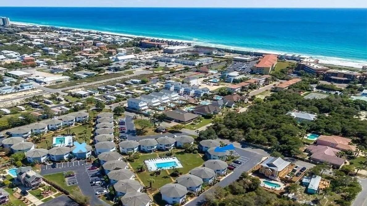 An aerial view of a coastal residential area with houses, a swimming pool, and a beach in the background.