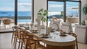 A dining area with a long table set for a meal, large windows offering a view of the sea and hills, and a cozy seating area with cushions and a plant. - Luxury, Seaview Villa Grandeca for 9+3 people (Rogoznica)