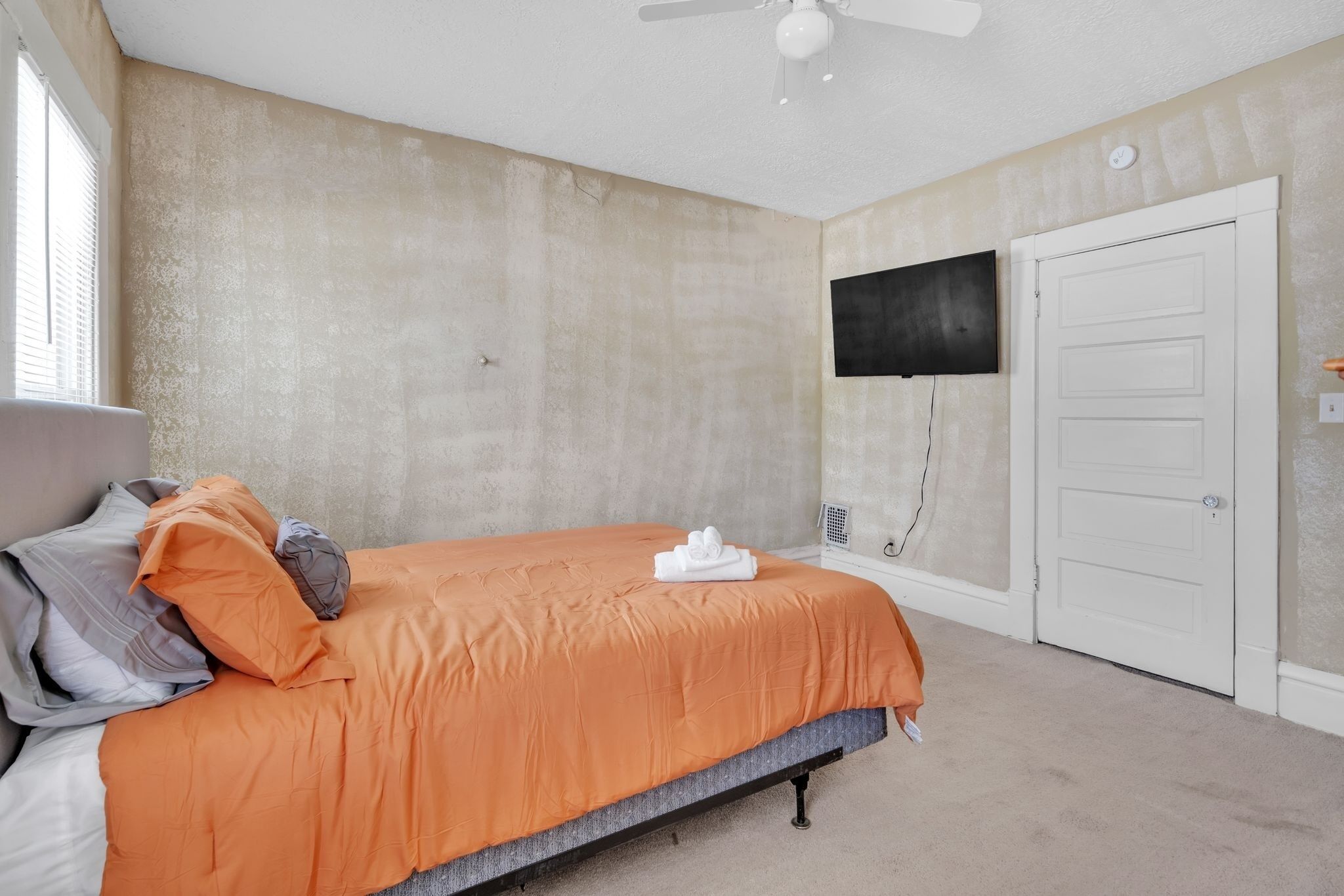 Une chambre à coucher avec un lit, un ventilateur de plafond, un téléviseur fixé au mur et une porte.