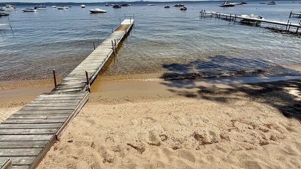 Una playa arenosa con un muelle de madera que se adentra en aguas tranquilas, barcos amarrados al fondo y un cielo despejado.