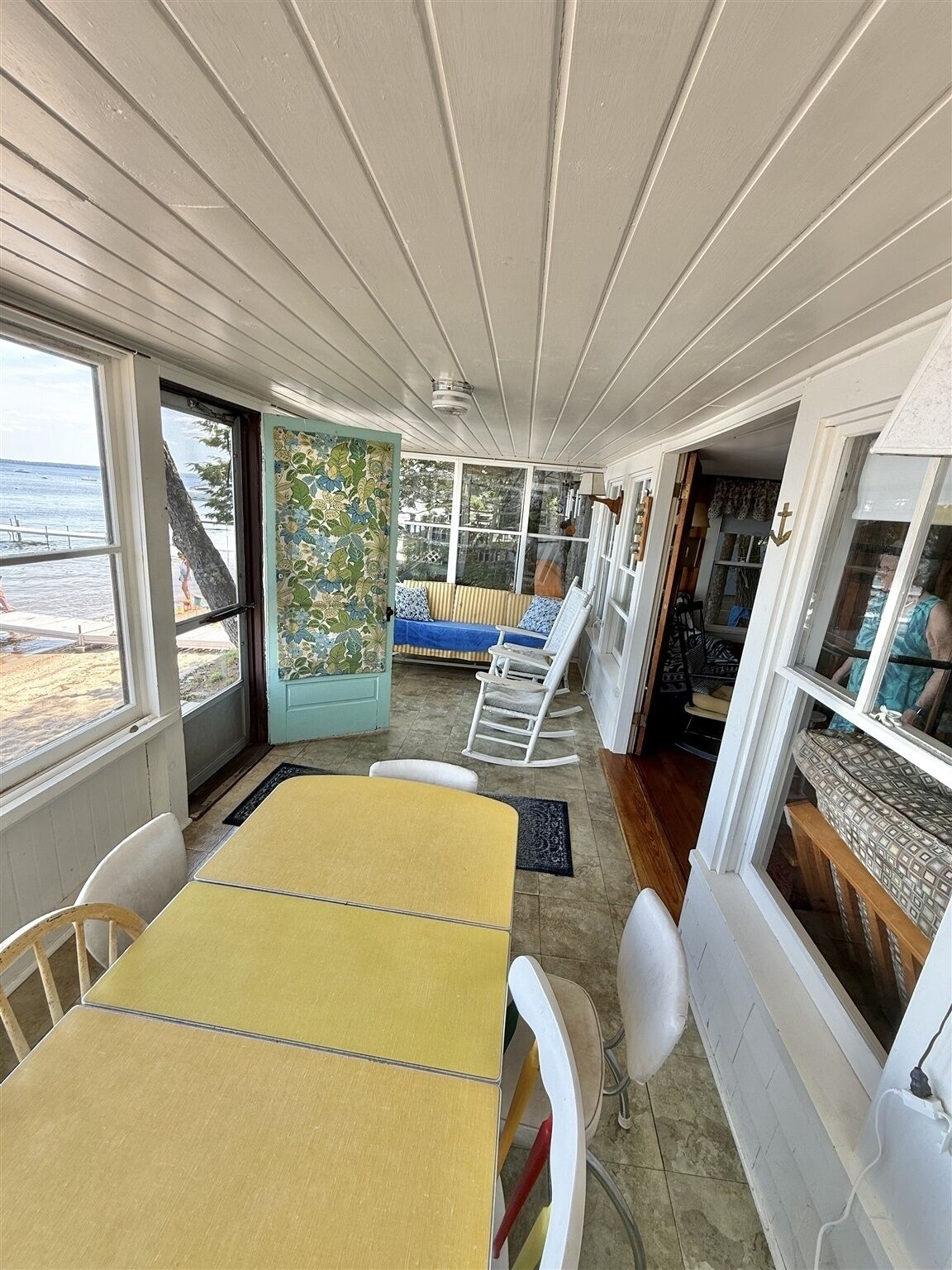 Une véranda avec une table jaune et des chaises blanches, un canapé bleu orné de coussins à motifs et une vue sur l’océan par de grandes fenêtres.