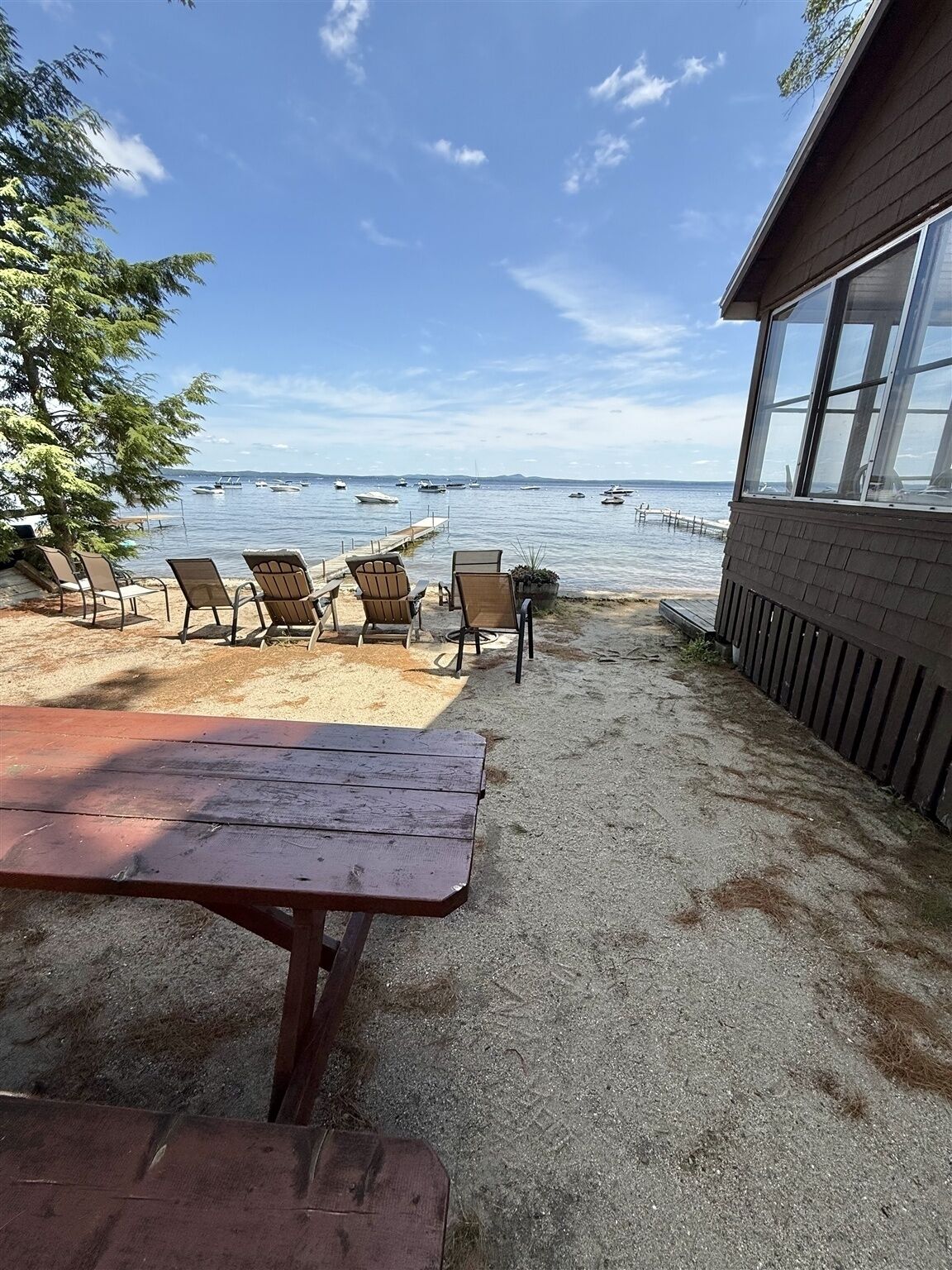 A beachside patio with a wooden table and several Adirondack chairs facing a calm sea with boats.