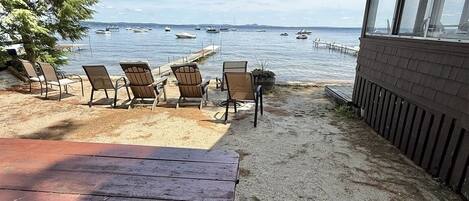 A beachside patio with a wooden table and several Adirondack chairs facing a calm sea with boats.