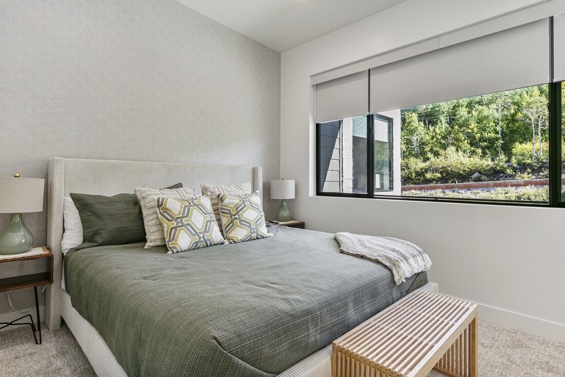 A bedroom with a large bed, a window with a view of trees, and a bedside table with a lamp.