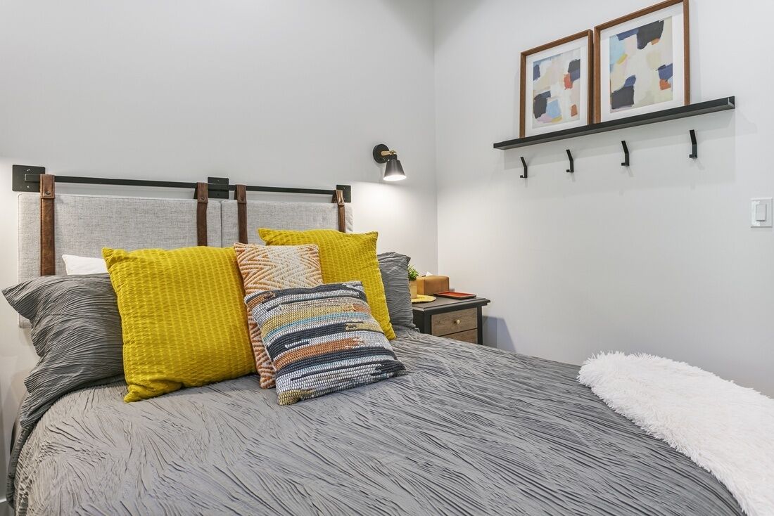 A neatly made bed with a grey textured bedspread and multiple pillows in various colors. There is a bedside table with a lamp and a small plant. The room has a white wall with a shelf holding two framed artworks.