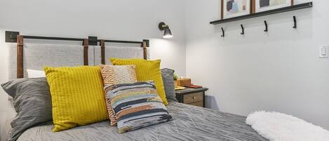 A neatly made bed with a grey textured bedspread and multiple pillows in various colors. There is a bedside table with a lamp and a small plant. The room has a white wall with a shelf holding two framed artworks.