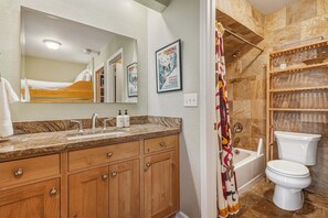 Une salle de bain avec un meuble-lavabo en bois, un grand miroir, une douche avec un rideau, des toilettes et une étagère contenant des articles de toilette.