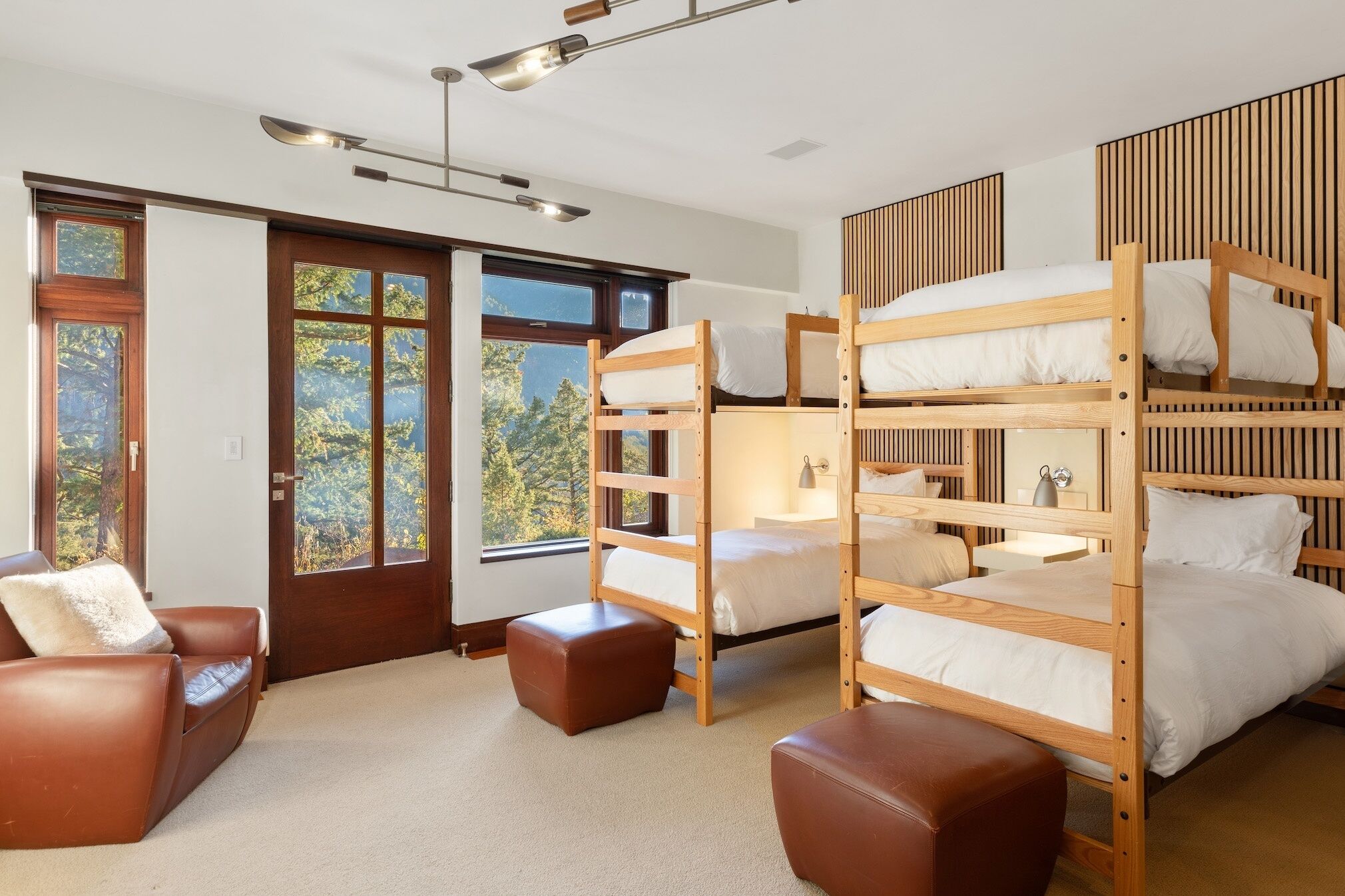 A bunk bed room with a brown leather sofa, a wooden desk, and a view of trees through the windows.