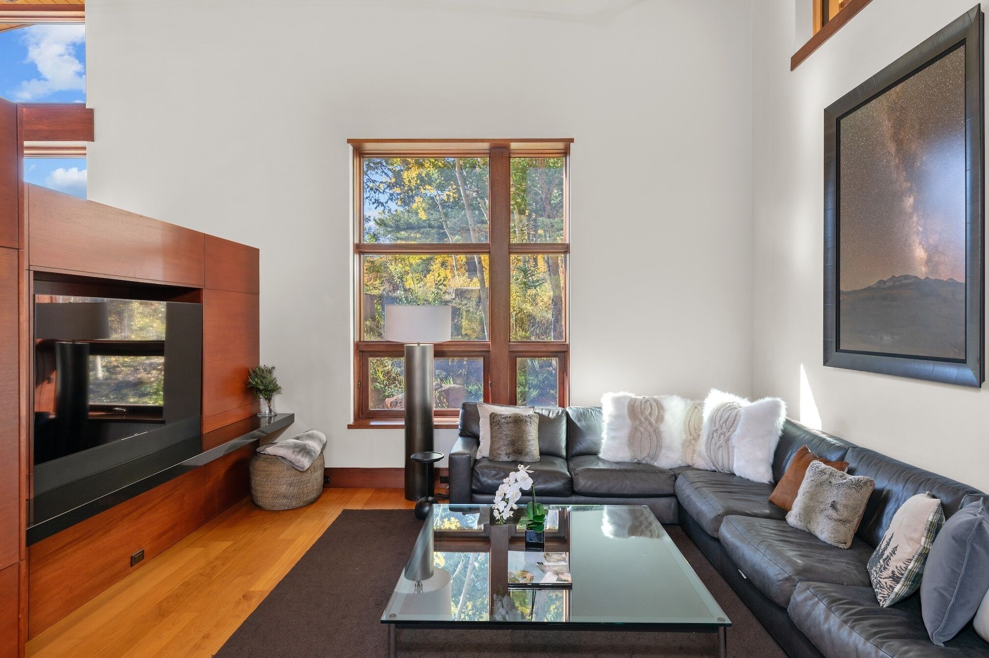 Una sala moderna con un sofá seccional, una mesa de centro de vidrio, una chimenea y ventanas amplias con vistas a árboles.