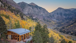 Eine Lodge mit einer großen Terrasse und zahlreichen Fenstern, eingebettet in eine gebirgige Landschaft mit Herbstlaub.