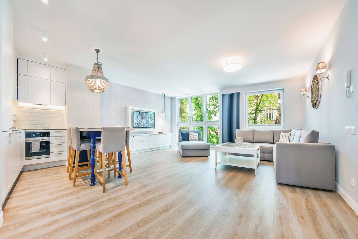 A modern kitchen with white cabinets, a dining area with a table and chairs, a gray sofa, and a large window with a view of greenery.
