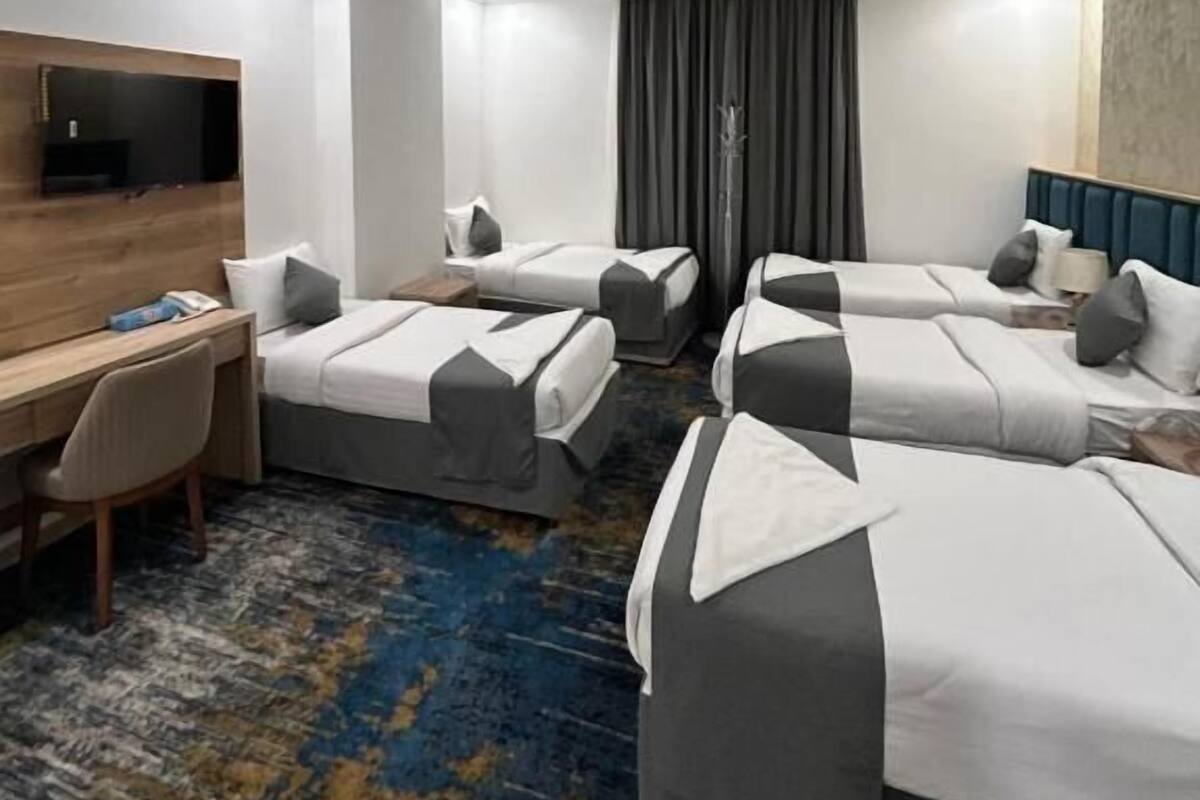 A hotel room with four beds arranged in a U-shape, a desk with a chair, a flat-screen TV mounted on the wall, and a patterned carpet.