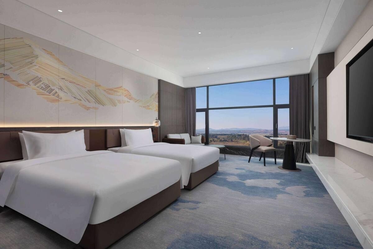 A hotel room with two beds, a large window with a view, a flat-screen TV, and a small table with chairs.