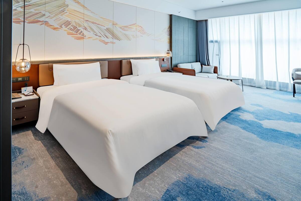 A hotel room with two beds, a desk with a lamp, a chair, and a large window with curtains.