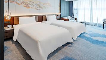 Habitación de hotel con dos camas, un escritorio con lámpara, una silla y un gran ventanal con cortinas.