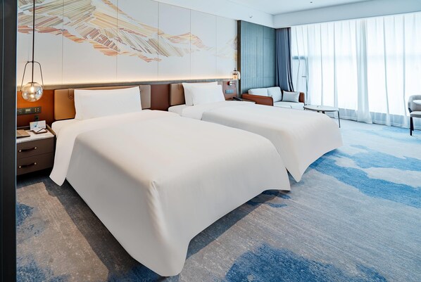 Habitación de hotel con dos camas, un escritorio con lámpara, una silla y un gran ventanal con cortinas.