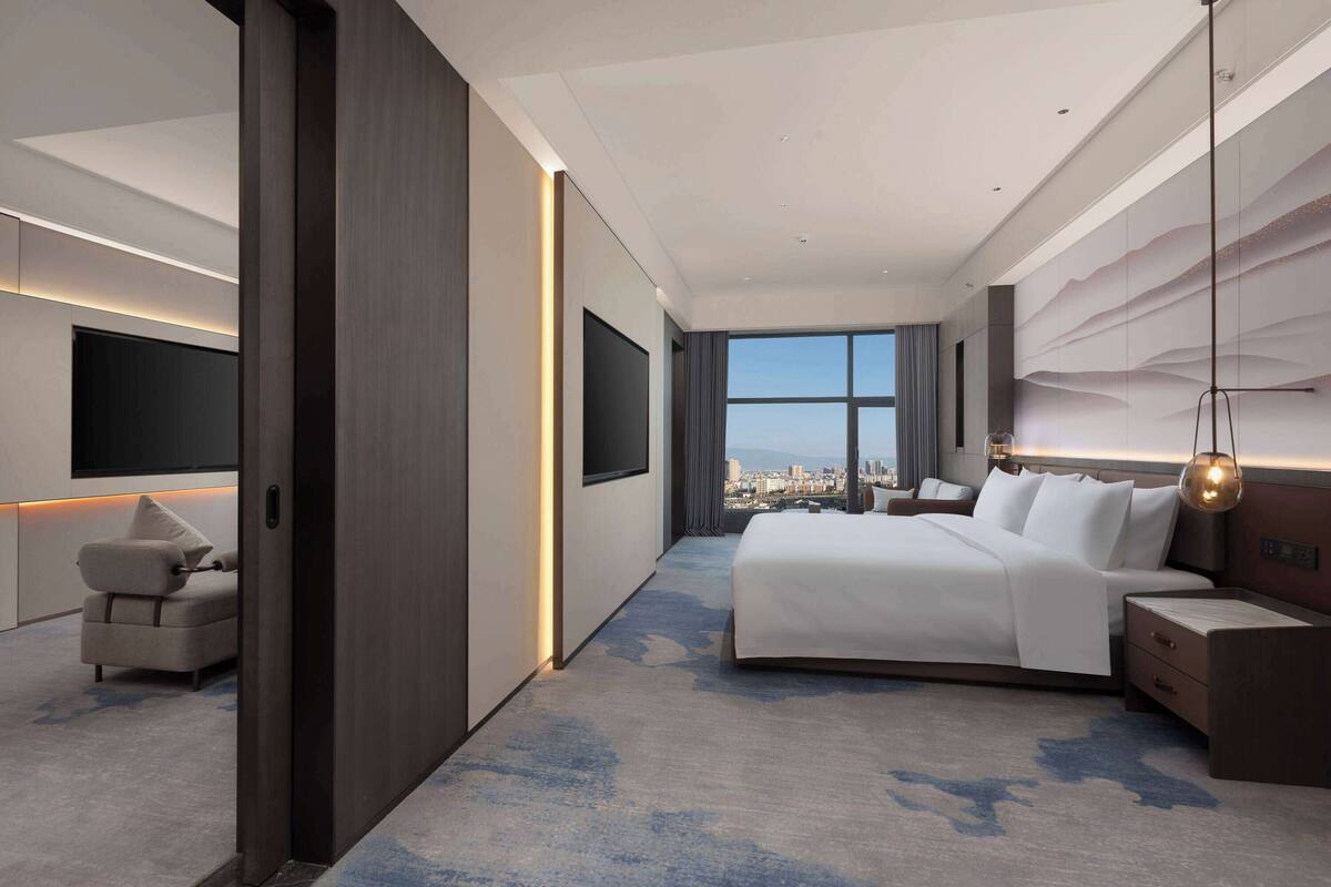 A modern hotel room with a large bed, a flat-screen TV mounted on the wall, a sofa, and a city view through the window.