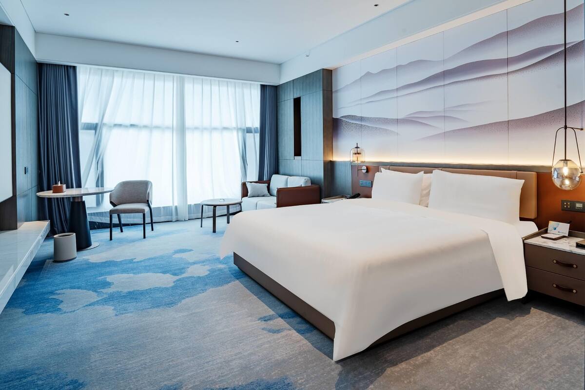 A modern hotel room with a large bed, a desk, a chair, and a view of the city through large windows.