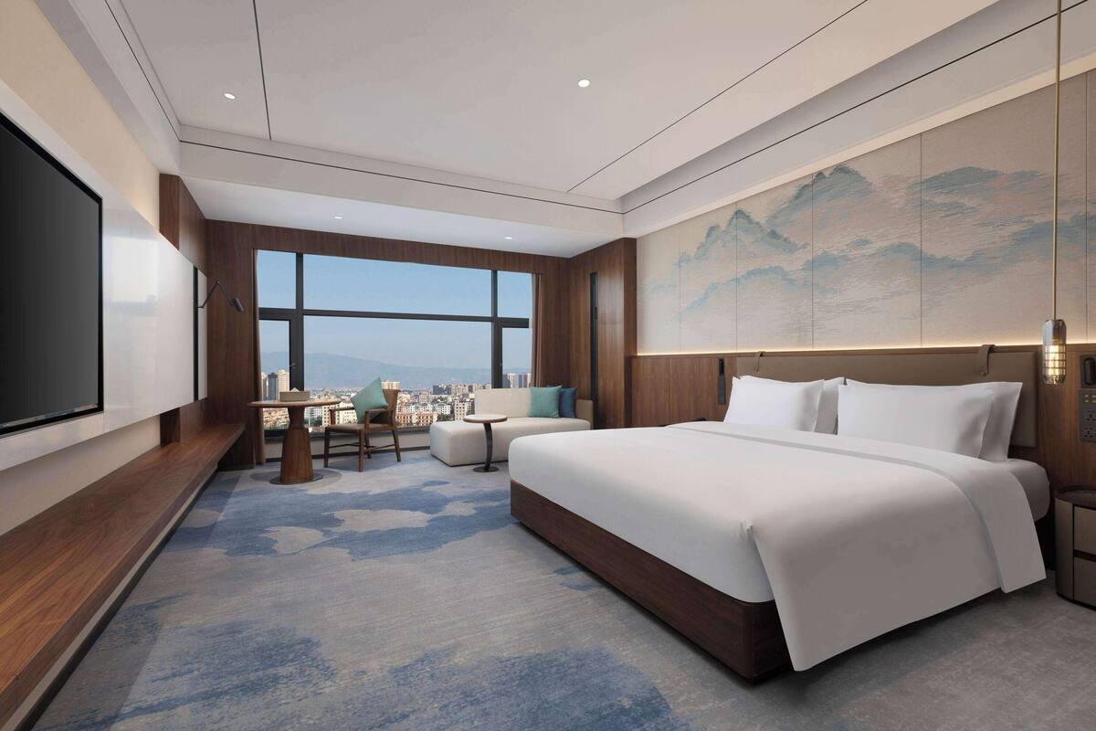 A modern hotel room with a large bed, a seating area with a table, and a city view through large windows.