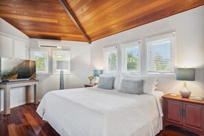 A bedroom with a wooden ceiling, white walls, a large bed, a TV on a stand, and a nightstand with a lamp. - Aloha Hale - Gorgeous home with pool, AC (Princeville)