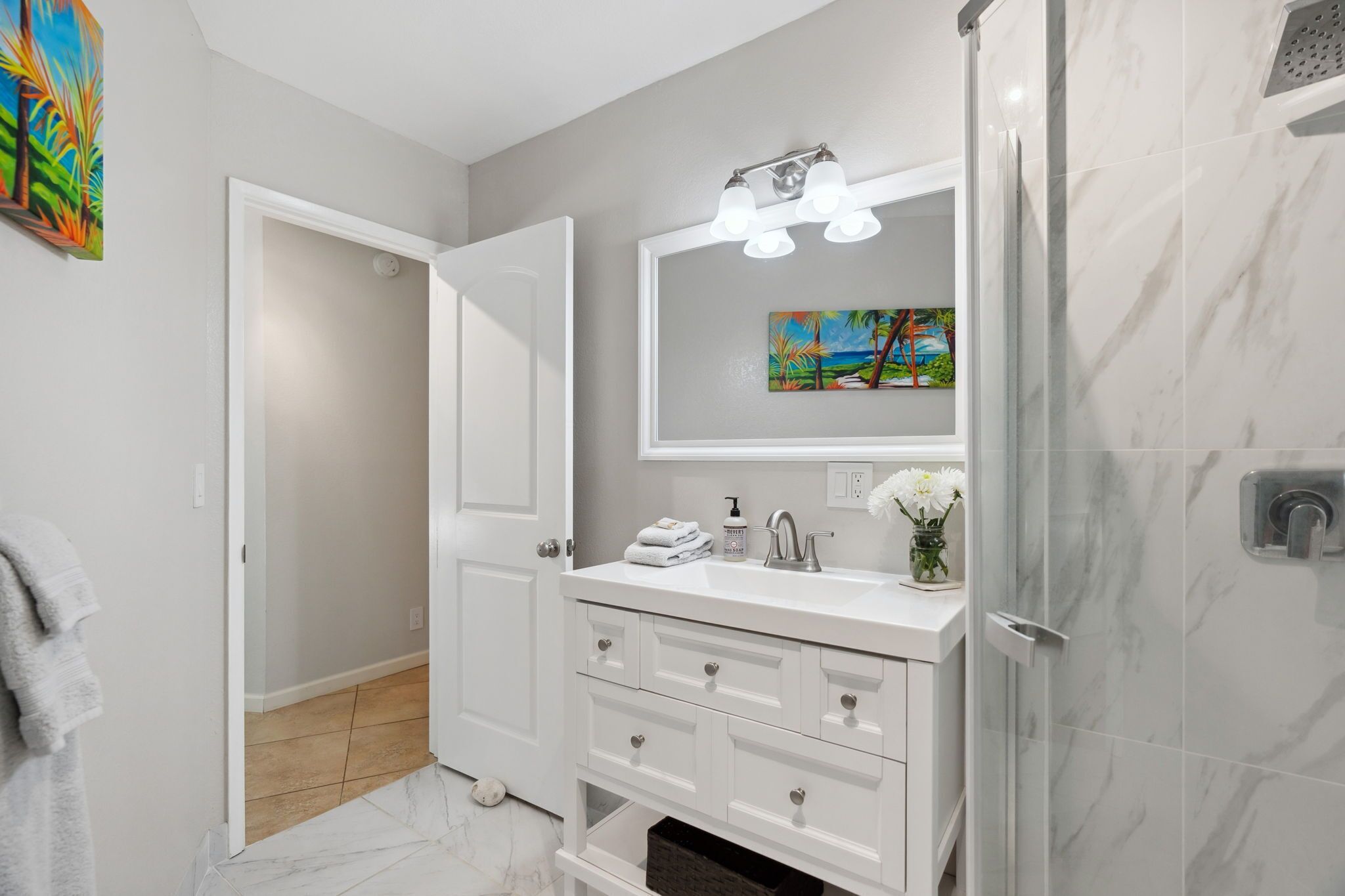 A modern bathroom with a white vanity, a shower with a glass door, and a painting above the mirror.