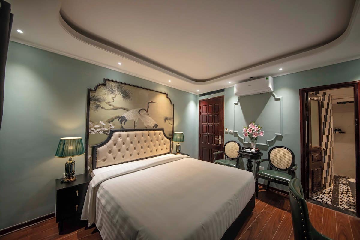 Een hotelkamer met een groot bed, een nachtkastje met lamp, een spiegel, een stoel en een deur naar een badkamer.