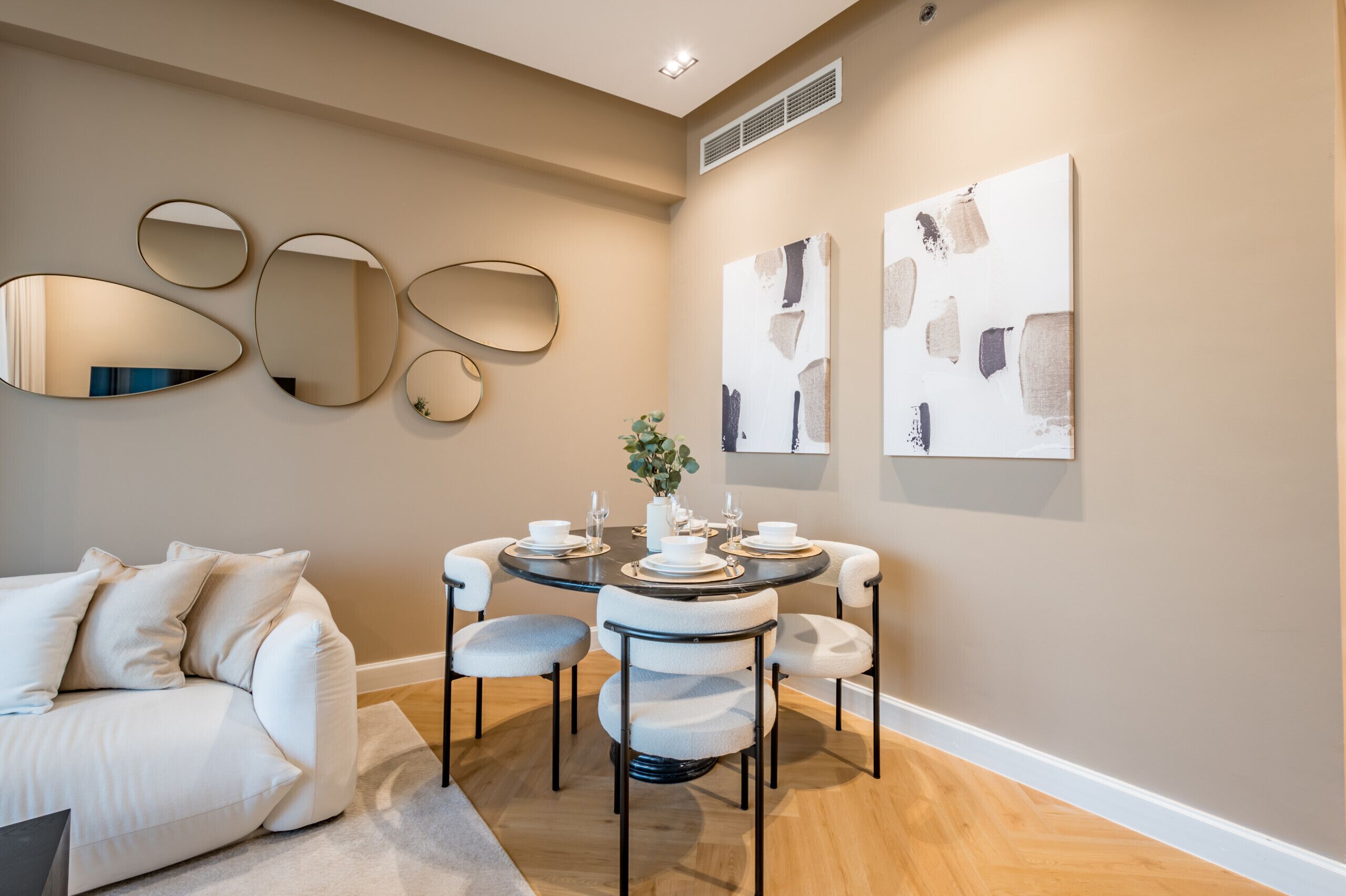 A modern dining area with a round table set for four, surrounded by chairs. There are abstract paintings on the wall and a series of oval mirrors. A white sofa is positioned in the foreground.