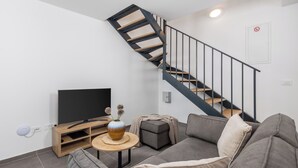 A modern living room with a grey sofa, a TV on a wooden stand, a vase with flowers, a small round table, and a staircase with a black railing. - Holiday Homes Oštro (Kraljevica)