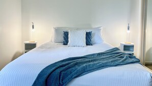 Una cama bien hecha con una manta azul, almohadas blancas y dos lámparas de noche sobre mesitas blancas.