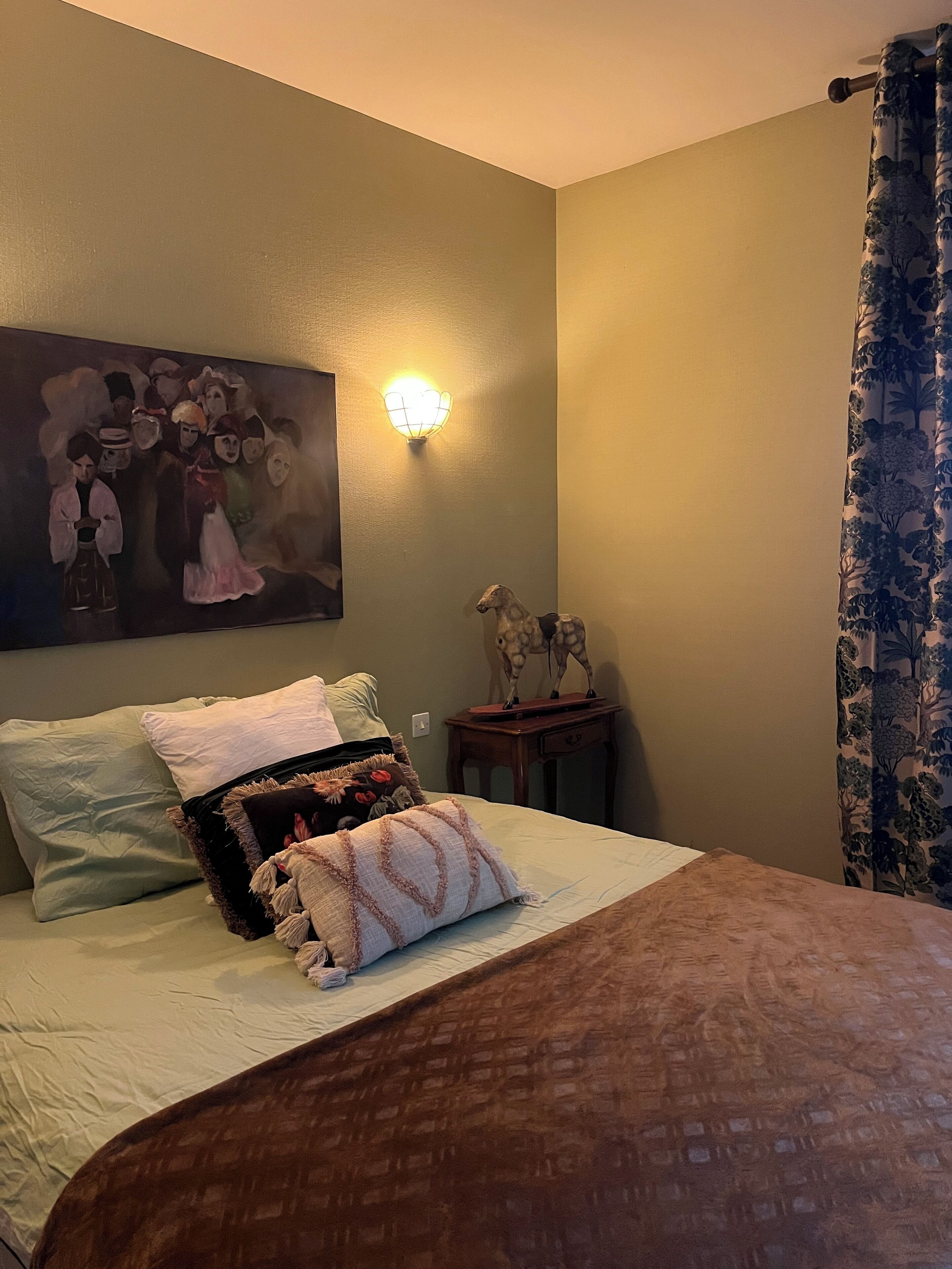 A bedroom with a bed, pillows, a wall-mounted light, a painting, and a small table with a statue.