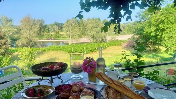 Tavolo apparecchiato all'aperto con vista su uno stagno e alberi, con croissant, frutta e una candela.