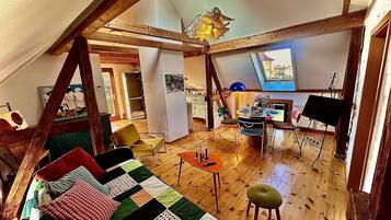 Ein gemütliches Loftzimmer mit Holzboden, einem farbenfrohen gesteppten Sofa, einem Esstisch mit Stühlen und einem Klavier.
