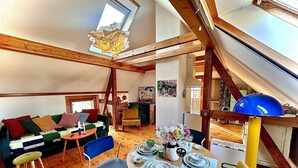 A cozy loft-style living space with a high ceiling, exposed wooden beams, a dining area with a table set for four, and a colorful sofa.