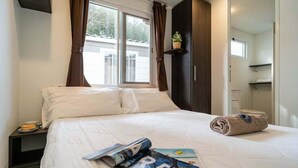 Una cama bien hecha con sábanas blancas, una ventana con cortinas, un estante con una planta y una toalla enrollada sobre la cama.