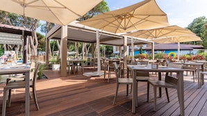Un espacio de patio al aire libre con piso de madera, mesas y sillas color beige bajo grandes sombrillas color beige.