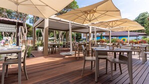 Un espacio de patio al aire libre con piso de madera, mesas y sillas color beige bajo grandes sombrillas color beige.